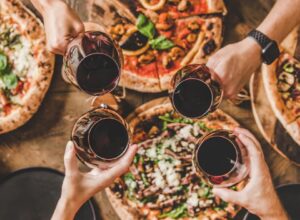 Image of friends with glasses of red wine cheering above pizza
