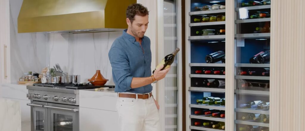 Image of a man in a kitchen holding a bottle of wine in front of a wine refrigerator